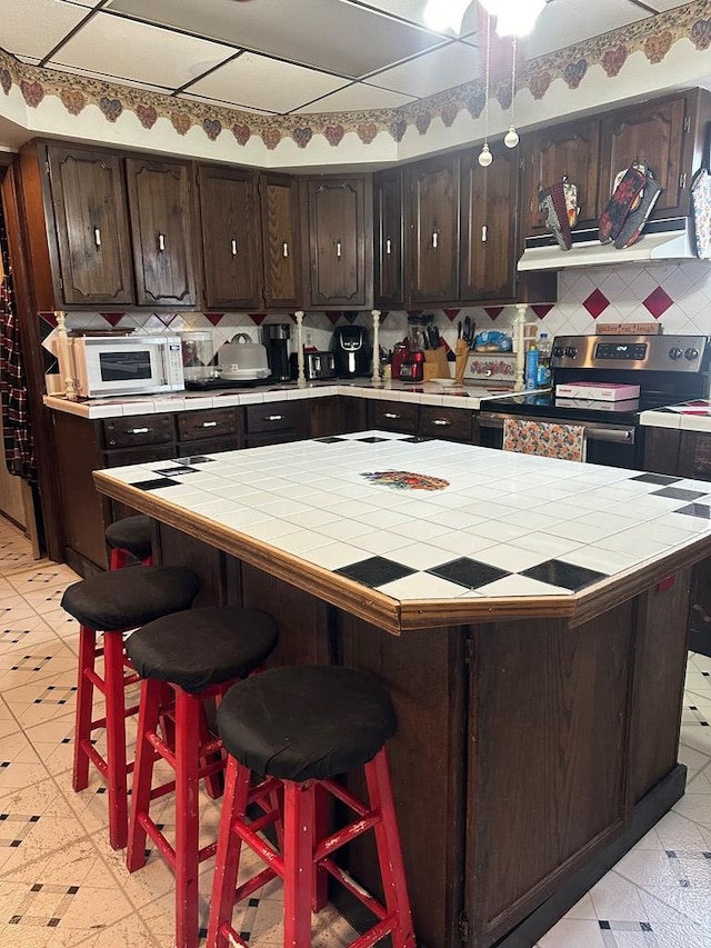 kitchen with backsplash, electric range, light tile patterned floors, dark brown cabinets, and a kitchen bar