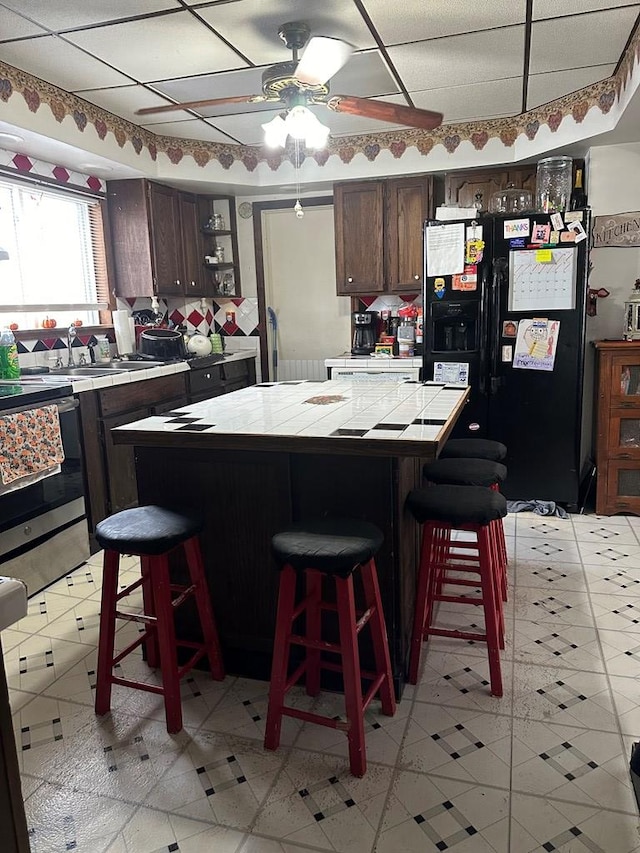 kitchen with a kitchen bar, black refrigerator with ice dispenser, dark brown cabinets, ceiling fan, and tile countertops