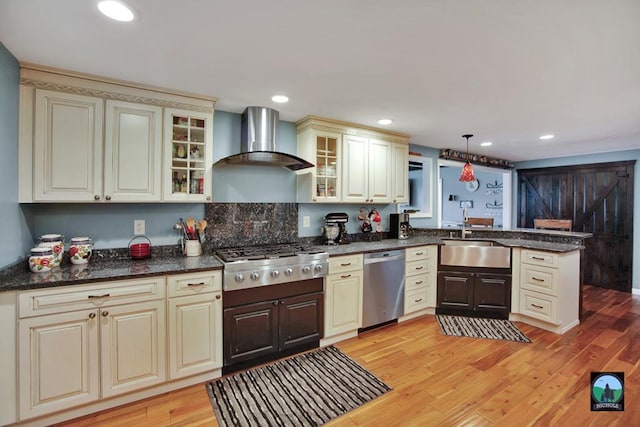 kitchen with wall chimney range hood, sink, hanging light fixtures, light hardwood / wood-style floors, and stainless steel appliances