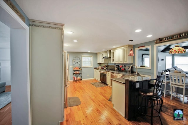 kitchen with wall chimney range hood, light hardwood / wood-style flooring, dark stone counters, pendant lighting, and a breakfast bar area