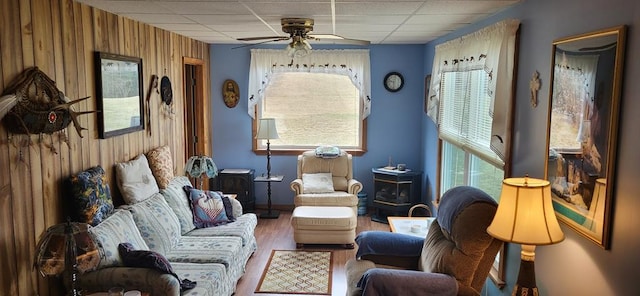 living area featuring wooden walls, wood finished floors, a wood stove, a drop ceiling, and ceiling fan