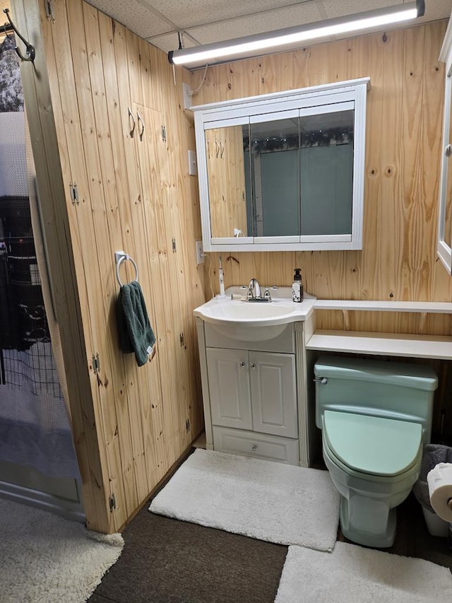 bathroom with toilet, vanity, wooden walls, and a drop ceiling