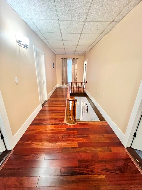 hall with wood-type flooring, baseboards, a drop ceiling, and an upstairs landing