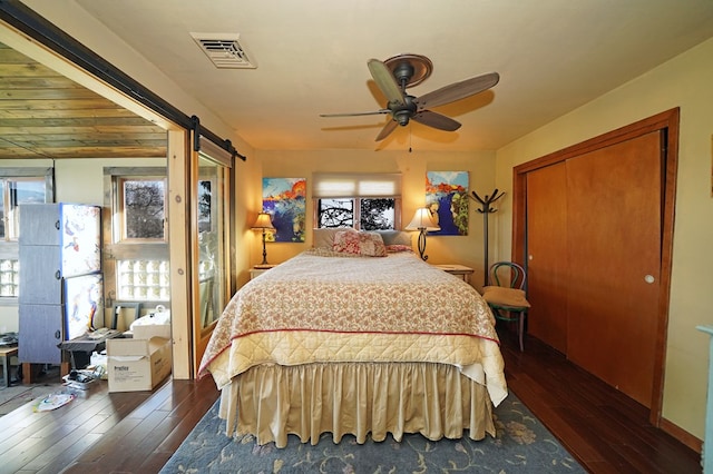 bedroom with a barn door, multiple windows, dark wood-type flooring, and ceiling fan