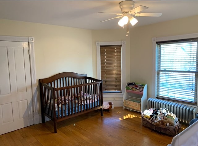 bedroom featuring radiator heating unit, hardwood / wood-style flooring, a nursery area, and ceiling fan