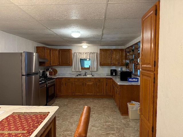 kitchen with brown cabinets, a sink, freestanding refrigerator, a drop ceiling, and gas range oven