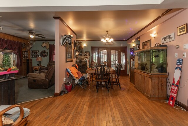 dining area with french doors, a stone fireplace, wood-type flooring, ceiling fan with notable chandelier, and ornamental molding