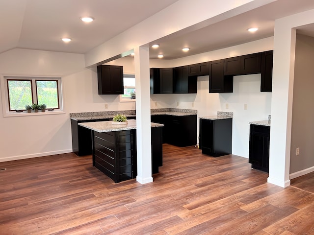 kitchen with wood finished floors, baseboards, light stone countertops, dark cabinets, and a center island