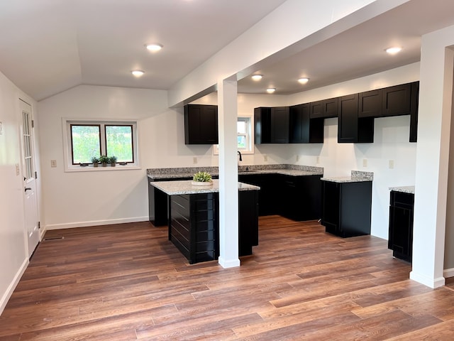 kitchen with light stone counters, a kitchen island, wood finished floors, dark cabinetry, and baseboards