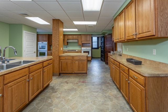 kitchen with under cabinet range hood, a drop ceiling, a sink, double oven, and brown cabinetry