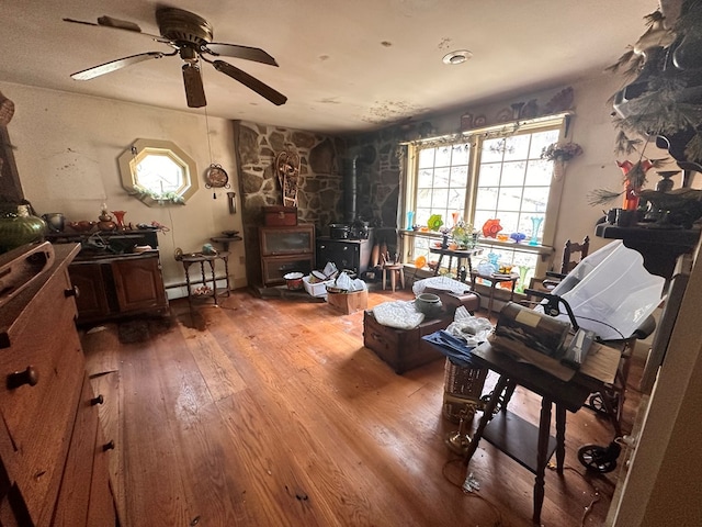 living area with a baseboard radiator, ceiling fan, a wood stove, and hardwood / wood-style floors