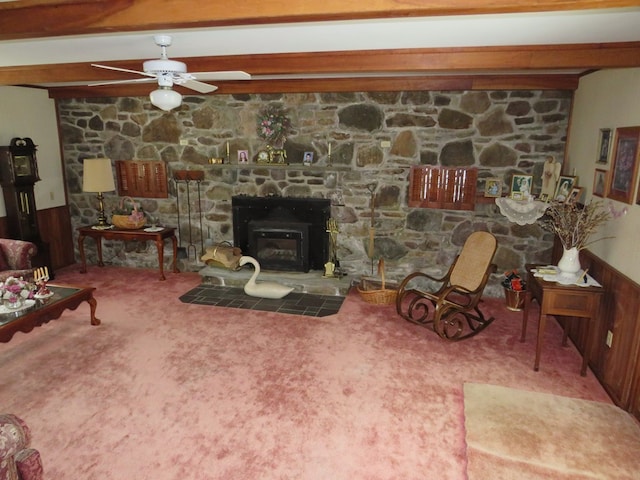 carpeted living room with beamed ceiling, ceiling fan, a stone fireplace, and wooden walls