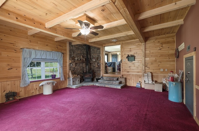 carpeted spare room featuring a wood stove, wood ceiling, wood walls, beamed ceiling, and ceiling fan