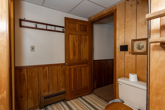 bathroom with a drop ceiling, wooden walls, toilet, and baseboard heating