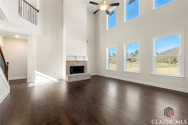 unfurnished living room with a towering ceiling, dark wood-type flooring, a brick fireplace, and ceiling fan