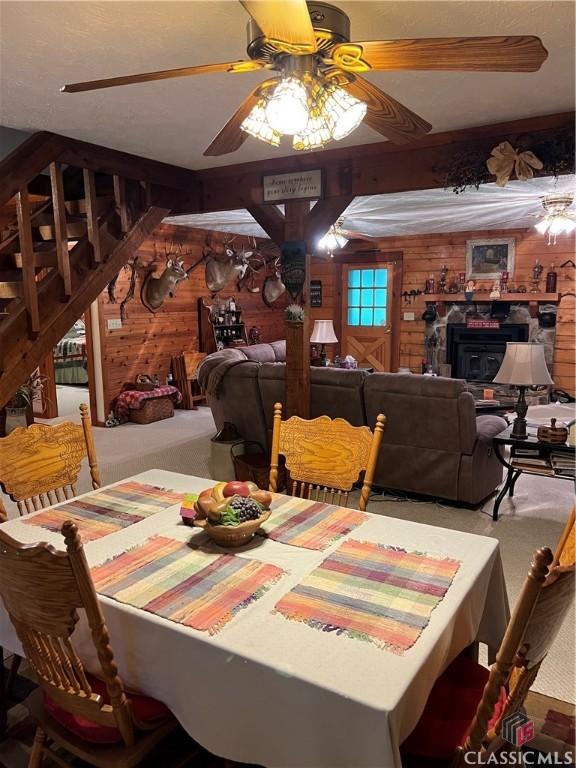 dining area with carpet, ceiling fan, wood walls, and a stone fireplace