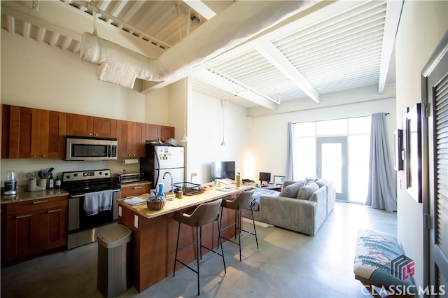 kitchen featuring a breakfast bar area, beam ceiling, a center island with sink, appliances with stainless steel finishes, and concrete floors