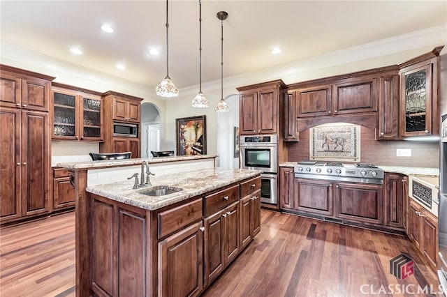 kitchen with a sink, stainless steel appliances, arched walkways, dark wood-style flooring, and a center island with sink