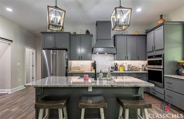 kitchen featuring dark wood-type flooring, premium range hood, stainless steel appliances, light stone countertops, and decorative light fixtures