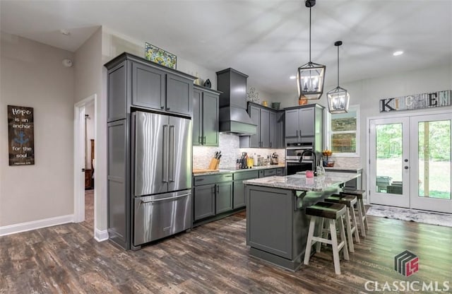 kitchen featuring light stone counters, hanging light fixtures, appliances with stainless steel finishes, an island with sink, and custom range hood