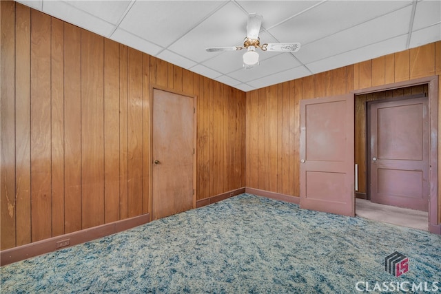 carpeted empty room with a paneled ceiling, ceiling fan, and wooden walls