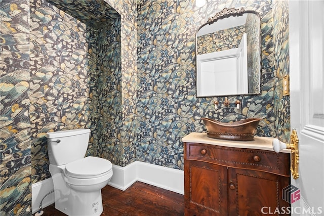 half bath with toilet, baseboards, wallpapered walls, wood finished floors, and vanity