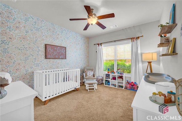 carpeted bedroom with a crib, wallpapered walls, ceiling fan, and visible vents