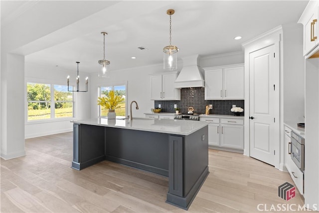 kitchen featuring premium range hood, sink, decorative light fixtures, white cabinetry, and an island with sink