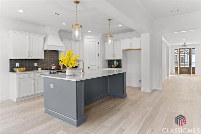 kitchen featuring premium range hood, backsplash, an island with sink, decorative light fixtures, and white cabinets