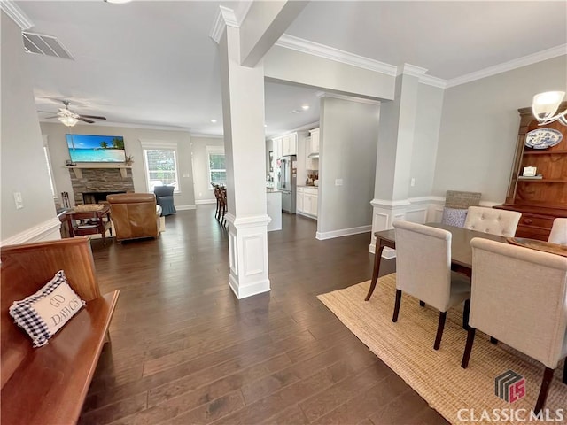 dining room featuring ornate columns, ornamental molding, ceiling fan, dark hardwood / wood-style floors, and a stone fireplace