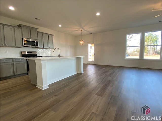 kitchen featuring a center island with sink, appliances with stainless steel finishes, gray cabinetry, decorative backsplash, and dark wood-type flooring