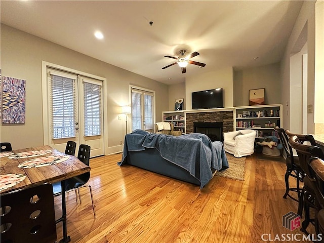 living room featuring ceiling fan, a stone fireplace, light hardwood / wood-style floors, and french doors