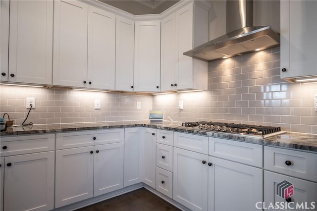 kitchen with wall chimney range hood, white cabinetry, dark stone countertops, stainless steel gas cooktop, and decorative backsplash