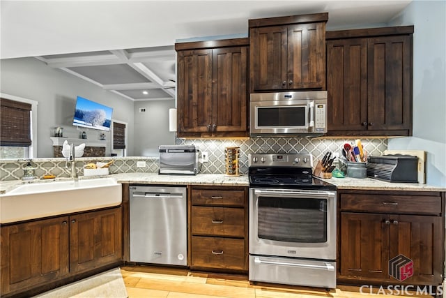 kitchen featuring dark brown cabinetry, coffered ceiling, sink, stainless steel appliances, and light stone countertops