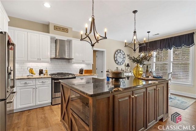 kitchen with hanging light fixtures, appliances with stainless steel finishes, a kitchen island, white cabinets, and wall chimney range hood