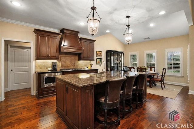 kitchen with dark hardwood / wood-style floors, decorative light fixtures, custom exhaust hood, dark stone counters, and a large island with sink