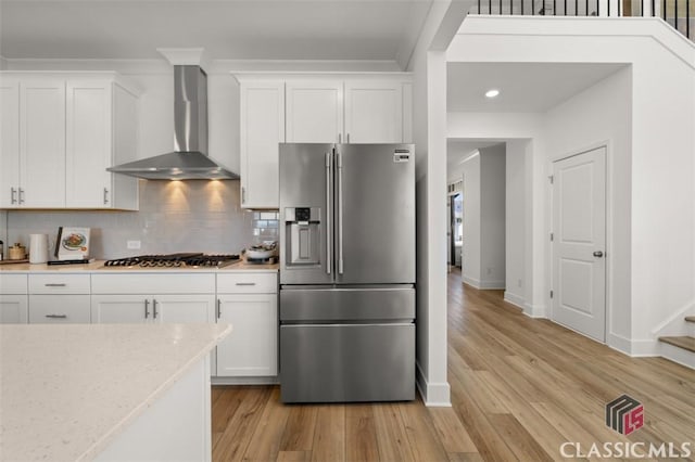 kitchen featuring wall chimney range hood, white cabinetry, stainless steel appliances, decorative backsplash, and light wood-type flooring