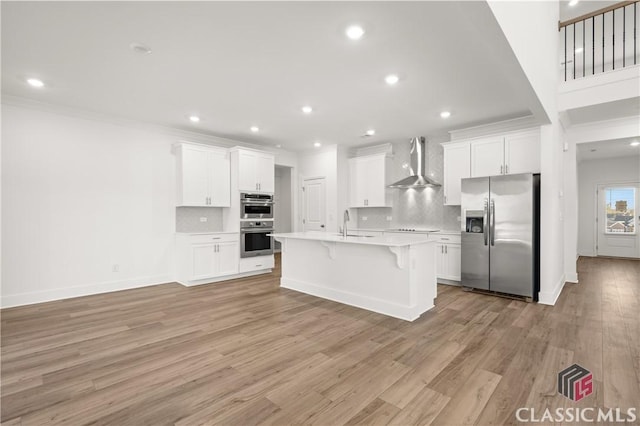 kitchen with wall chimney exhaust hood, appliances with stainless steel finishes, a kitchen island with sink, and white cabinets