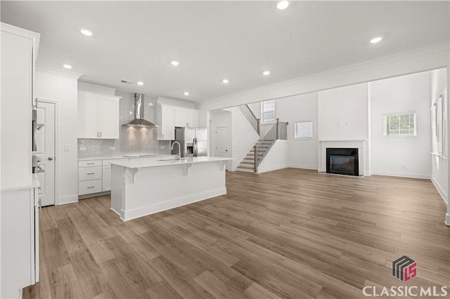 kitchen with stainless steel refrigerator with ice dispenser, a breakfast bar area, white cabinetry, a kitchen island with sink, and wall chimney range hood