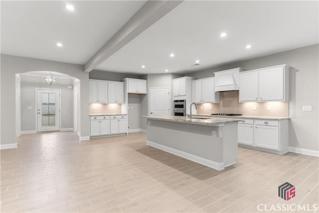 kitchen featuring sink, double oven, light stone counters, an island with sink, and white cabinets