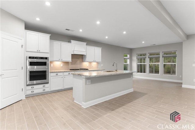 kitchen featuring double oven, sink, white cabinets, light stone countertops, and a center island with sink