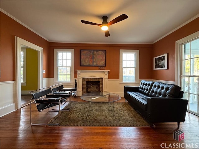 living area with a brick fireplace, crown molding, ceiling fan, wainscoting, and wood finished floors