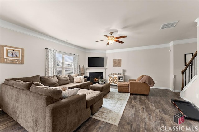 living area with crown molding, dark wood finished floors, visible vents, stairway, and a warm lit fireplace