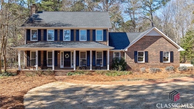 colonial home featuring covered porch, roof with shingles, a chimney, and brick siding