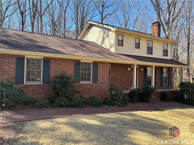 view of front of house featuring brick siding, a chimney, a shingled roof, covered porch, and a front lawn