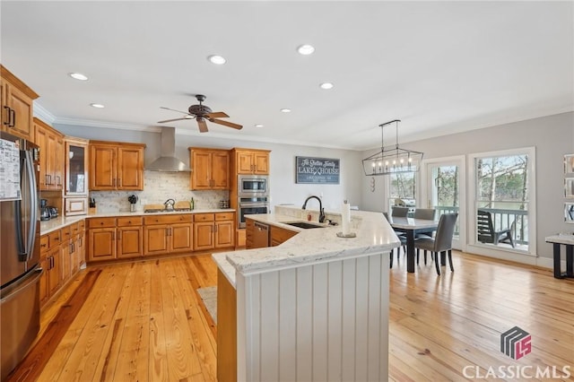 kitchen with appliances with stainless steel finishes, brown cabinetry, a kitchen island with sink, a sink, and wall chimney exhaust hood