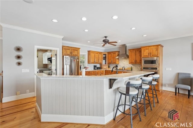 kitchen featuring brown cabinetry, appliances with stainless steel finishes, a kitchen breakfast bar, a large island with sink, and wall chimney range hood