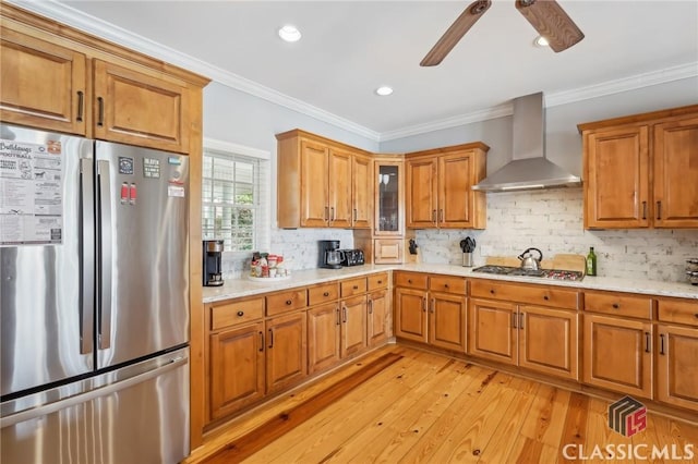 kitchen with light wood finished floors, wall chimney exhaust hood, ornamental molding, stainless steel appliances, and backsplash