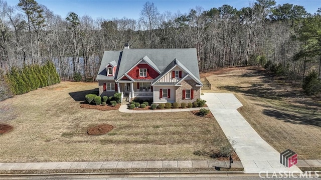 view of front facade with a porch, concrete driveway, a front yard, stone siding, and a forest view