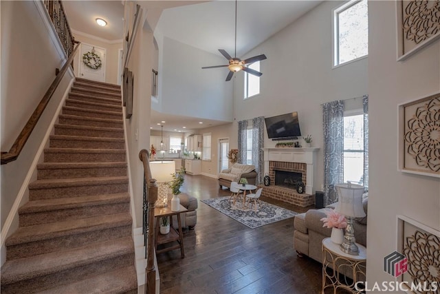 living room featuring a ceiling fan, baseboards, stairs, a brick fireplace, and dark wood-style floors